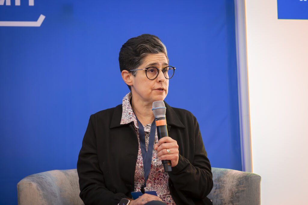 Margo Martinez is sitting in a chair, wearing a black blazer with a floral undershirt. Her hair dark, and cropped shortly, slightly graying on the left side. She has round glasses and holds a microphone up as she speaks. 