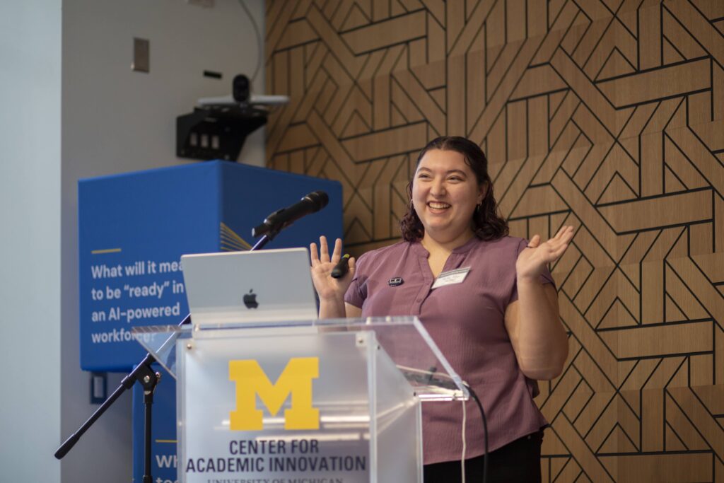 Person smiling and gesturing with both hands while presenting at a podium with a microphone and laptop.