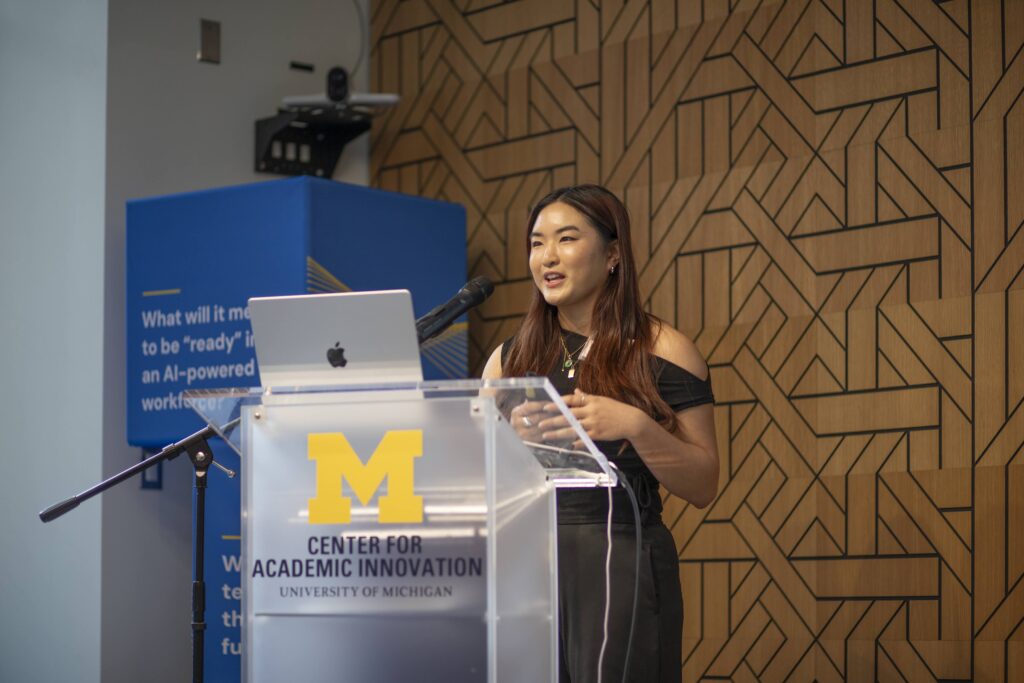 A woman stands at a clear podium with a microphone and open laptop, speaking while holding a small remote.