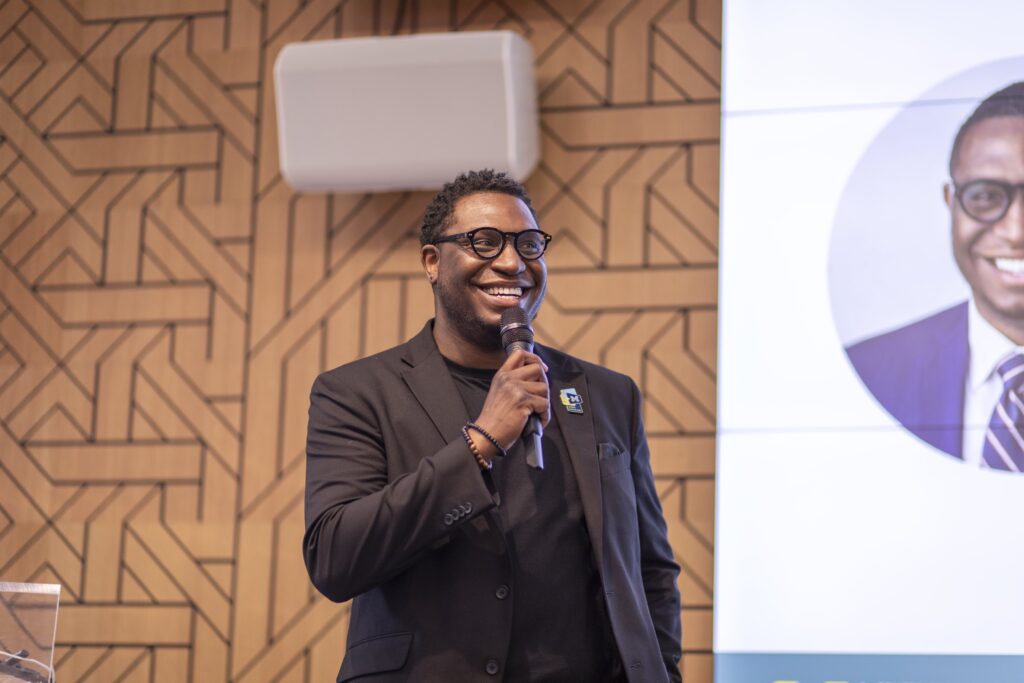 A man wearing glasses and a black blazer smiles while holding a microphone and speaking.