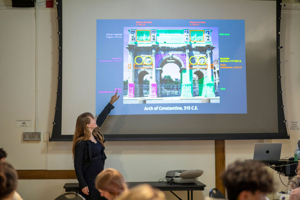 A woman stands at the front of a classroom pointing to a projected slide about the Arch of Constantine while students sit in the foreground.
