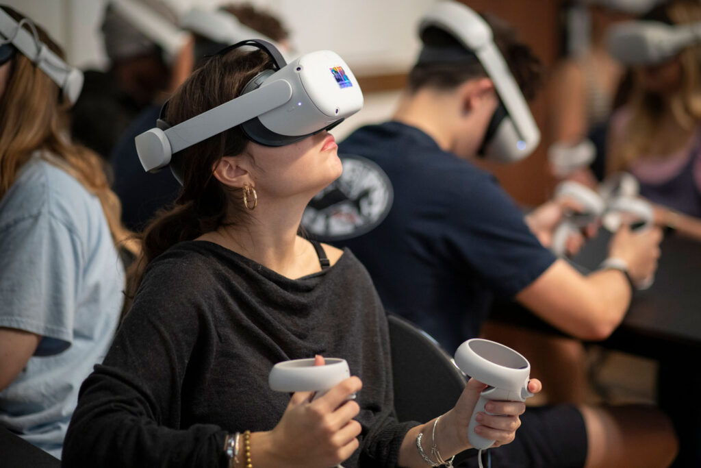 A student wearing a virtual reality headset holds two controllers during an immersive classroom activity.