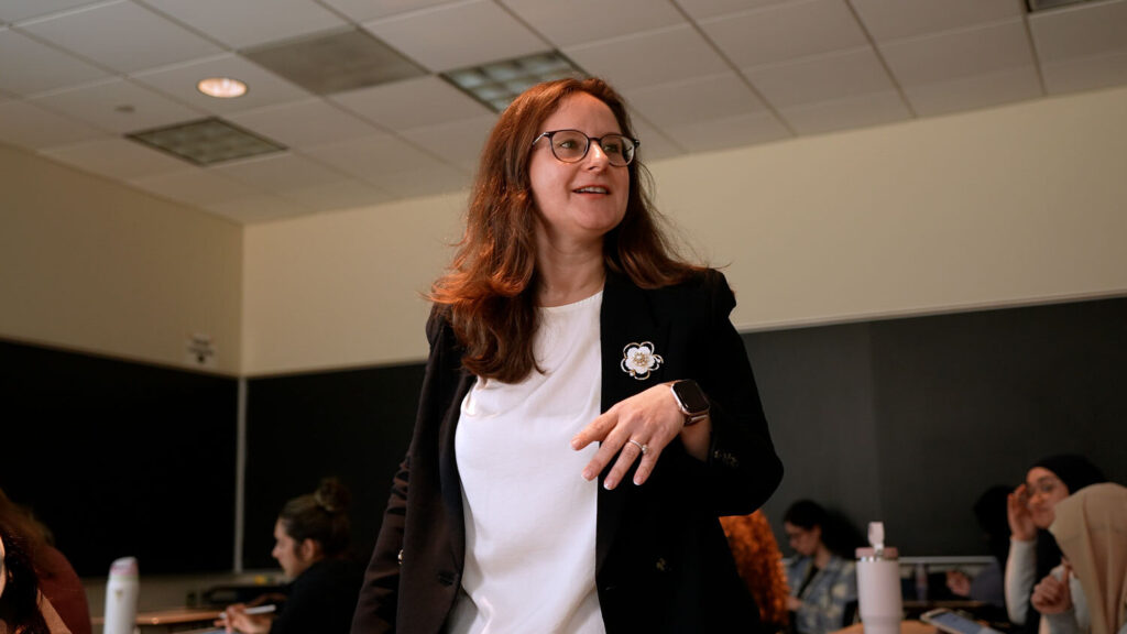 Woman stands smiling in middle of classroom, giving a lecture to her students.