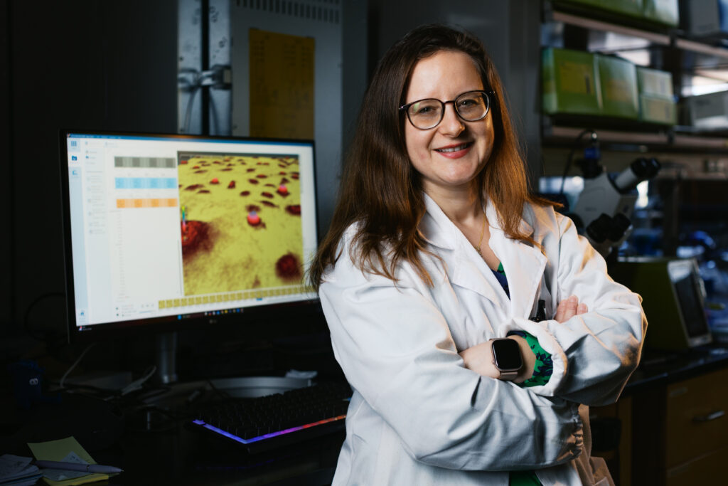 Woman stands smiling with her arms crossed in lab coat in front of computer.