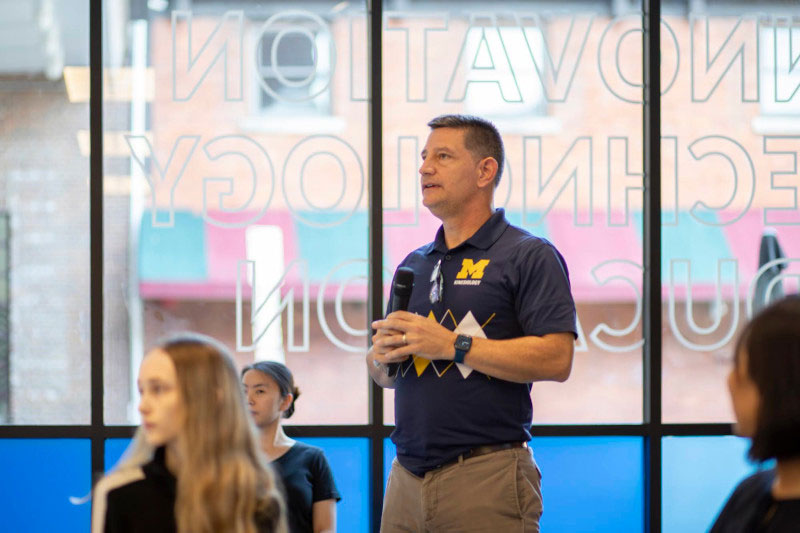 Man in a University of Michigan Kinesiology shirt stands holding a microphone. 
