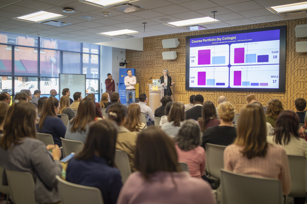 A woman speaks in front of a podium while two co-presenters stand off to the side. A large crowd watches the speaker and the slides on a big screen. 