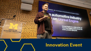 A speaker presents onstage at the University of Michigan Center for Academic Innovation, with a large screen behind him displaying a presentation about Unreal Engine and the automotive industry.