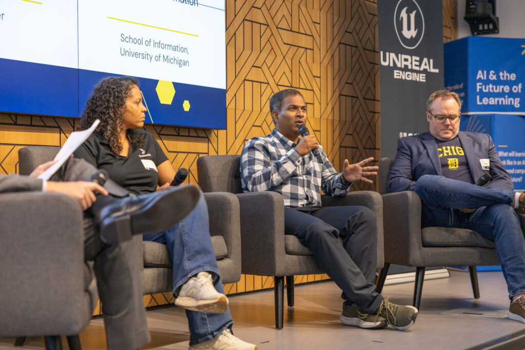 Three panelists sit onstage as the center speaker talks into a microphone, with University of Michigan and Unreal Engine branding behind them.
