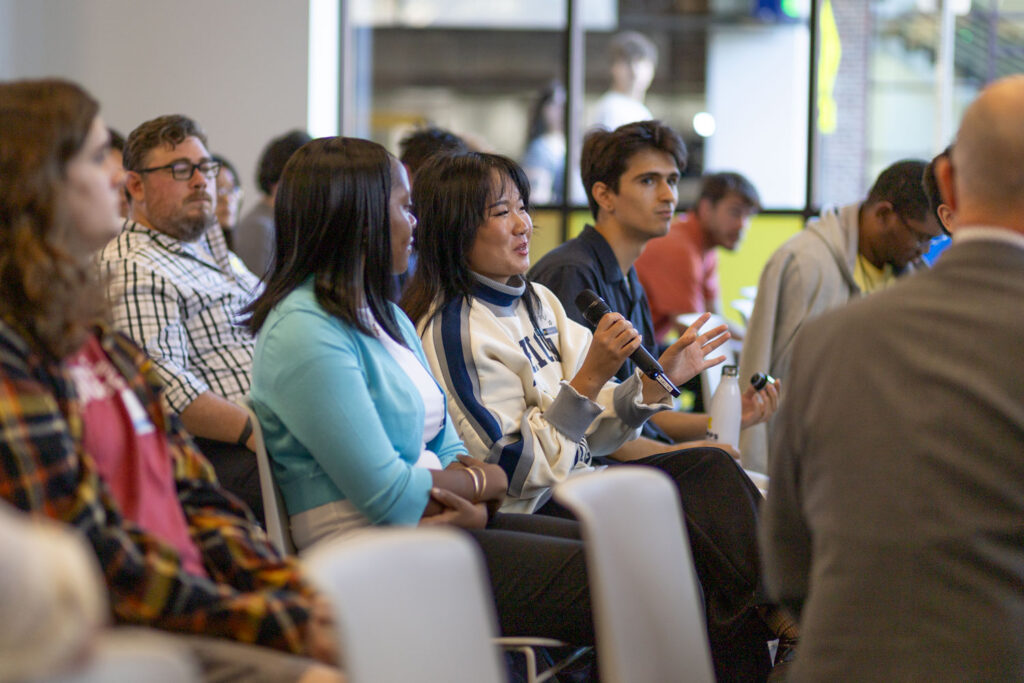 An audience member speaks into a handheld microphone while seated among other attendees at an event.
