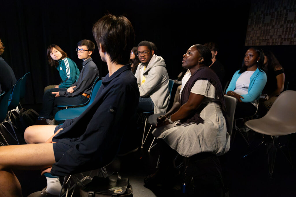 A group of students and other attendees sit in rows of chairs, watching a presentation in a darkened event space.