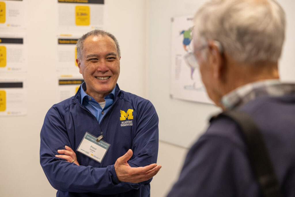 A man wearing a navy University of Michigan Academic Innovation jacket and a name badge reading Peter smiles and gestures while conversing with an older man. Posters and diagrams displayed on the wall behind them suggest an academic showcase or networking event.