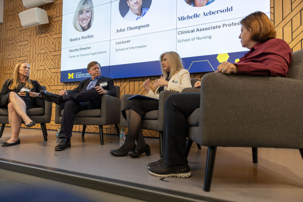 Four panelists sit in gray armchairs onstage during a University of Michigan event. A woman in a white blazer speaks while gesturing with her hands, as the other three panelists listen. A large screen behind them displays faculty portraits, names, and university affiliations.