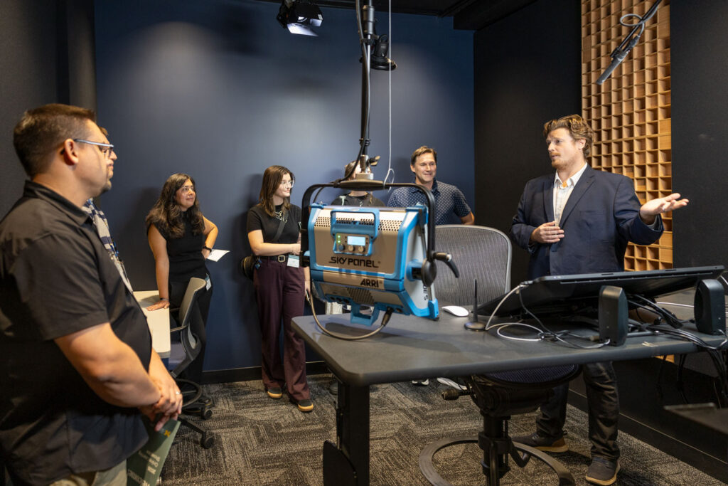 A man in a navy blazer speaks to a small group of attendees inside a recording studio, gesturing toward the equipment. The group stands around a table holding a large studio light, monitors, and audio equipment, with overhead microphones and soundproofing panels visible.