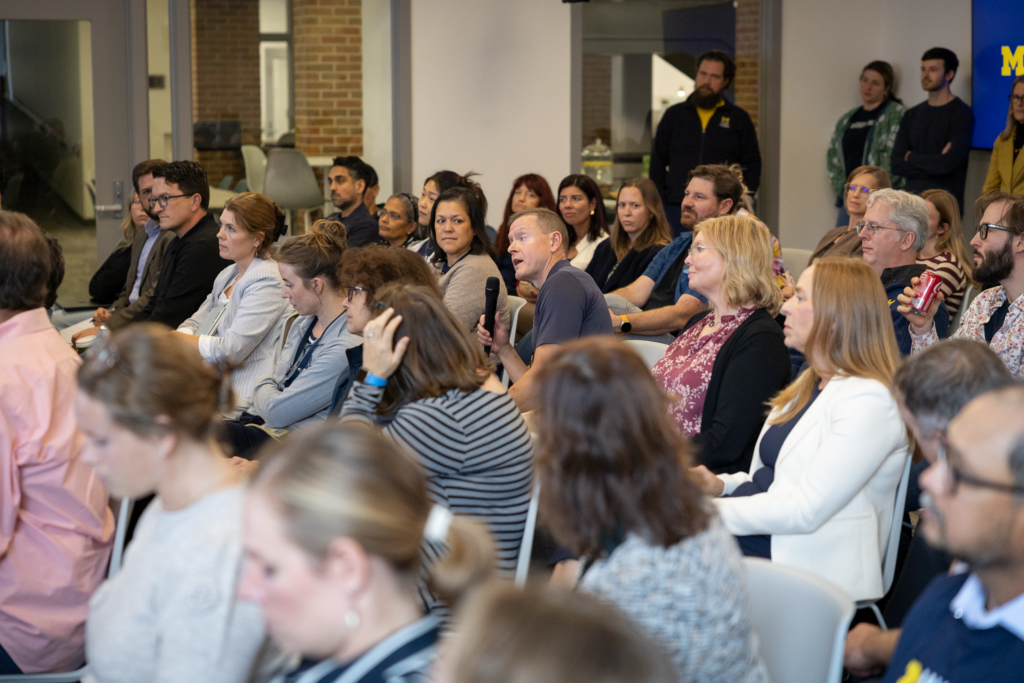 A man in a navy shirt holds a microphone and speaks from his seat during an audience discussion. Attendees seated around him listen attentively in a modern event space with glass walls and exposed brick.