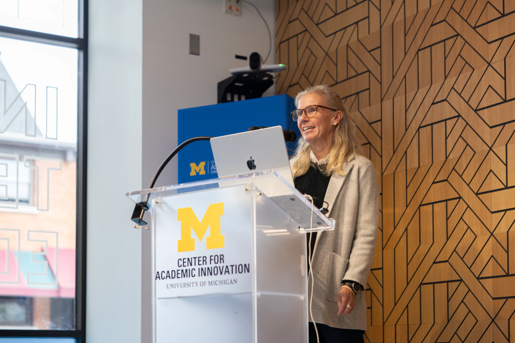 A woman with long blonde hair and glasses speaks into a microphone behind a clear podium displaying the University of Michigan Center for Academic Innovation logo. An open laptop sits on the podium, with a geometric wood-paneled wall behind her.