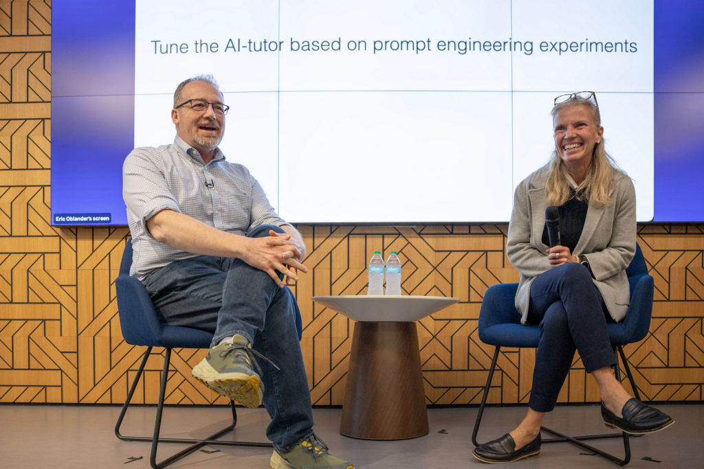 Two speakers sit onstage at an Innovation Event. A man in a light blue shirt gestures while speaking, and a woman with long blonde hair holds a microphone and smiles. Behind them, a screen displays the text, "Tune the AI-tutor based on prompt engineering experiments."