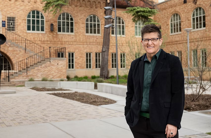 Person wearing a blazer and glasses stands in courtyard. 